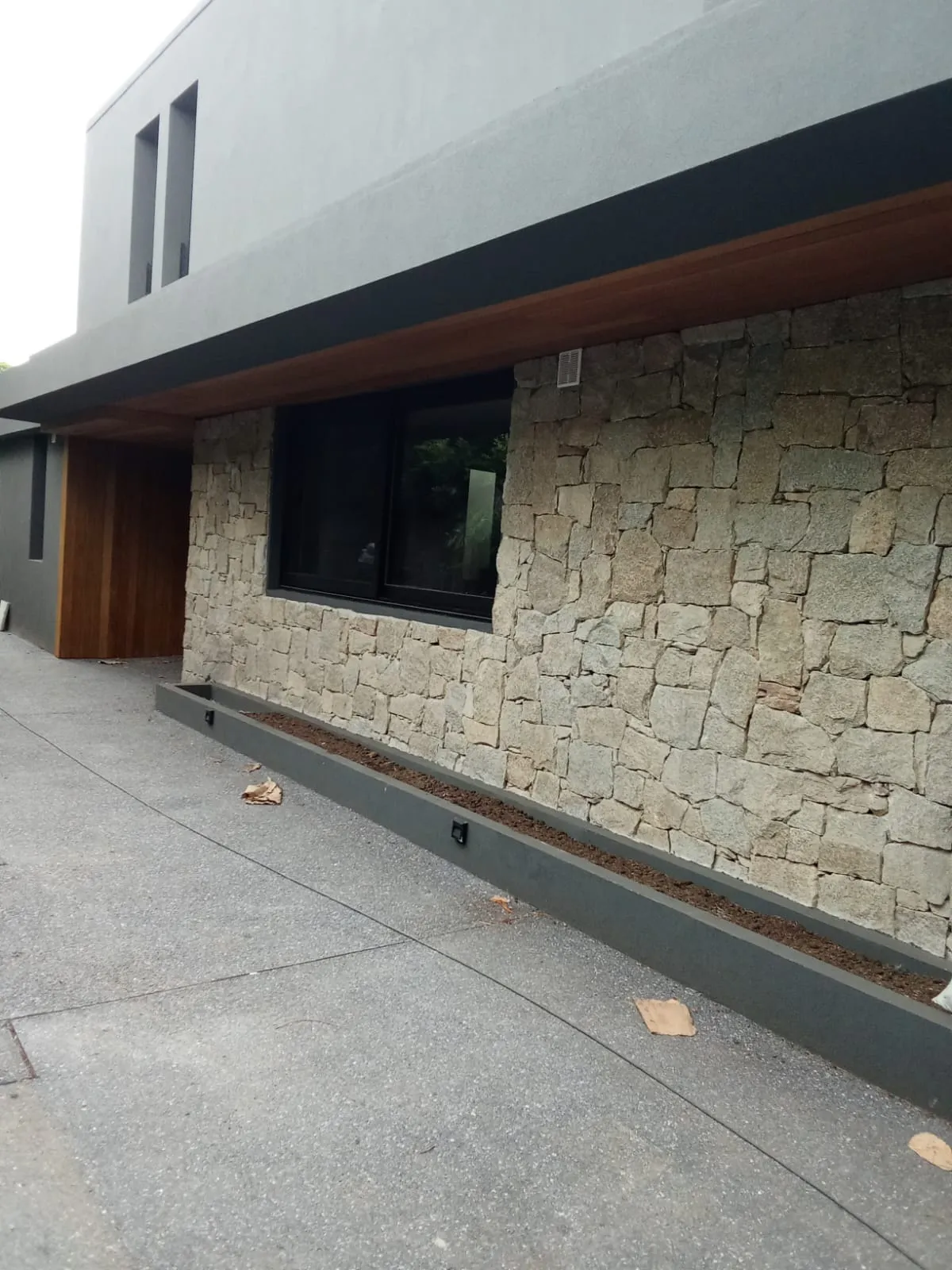 Fachada con muro de piedra natural, alero de madera y ventana negra